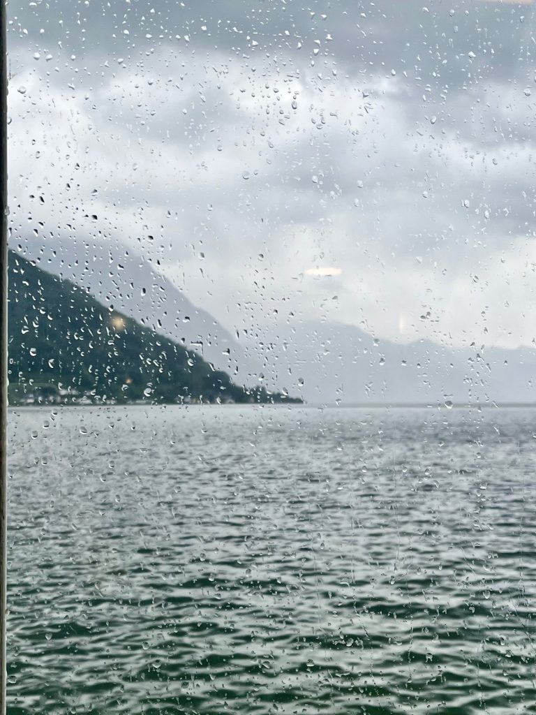 Regentropfen auf einer Fensterscheibe mit Blick auf einen See und bewölkte Berge