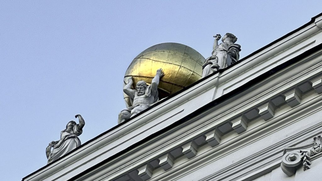 Atlas-Statue auf einem Wiener Barockgebäude, die einen goldenen Globus auf den Schultern trägt, vor hellblauem Himmel.