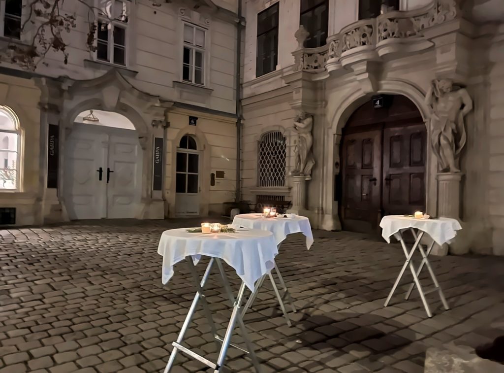 Barocker Innenhof bei Nacht mit weiß gedeckten Stehtischen und Kerzen, beleuchtete Torbögen und Statuen im Hintergrund.