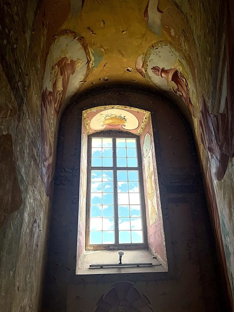 Freskos rund um das Fenster, dadurch sieht man blaue Wolkenam hellblauen Himmel.